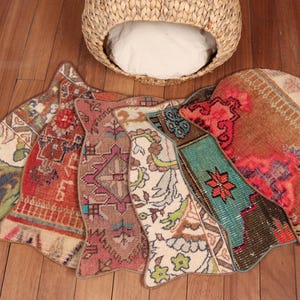 May include: A collection of colorful, patterned rugs in various shapes and sizes, arranged on a wooden floor. The rugs feature intricate designs in shades of red, blue, green, and brown. A woven basket with a white cushion is in the background.