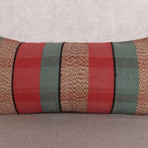 May include: Rectangular decorative pillow with a striped pattern. The pillow features alternating bands of red, green, and tan, with black accent lines. The pillow is placed on a neutral surface, suggesting a home decor item.