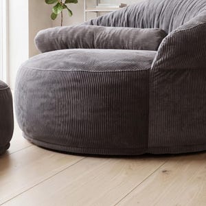May include: A dark gray corduroy armchair featuring a rounded, low-profile silhouette with a thick, horizontal bolster cushion across the backrest. The fabric has a distinct vertical ribbed texture, and the chair has a deep, wide seat design.
