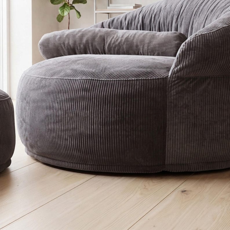 May include: A dark gray corduroy armchair featuring a rounded, low-profile silhouette with a thick, horizontal bolster cushion across the backrest. The fabric has a distinct vertical ribbed texture, and the chair has a deep, wide seat design.