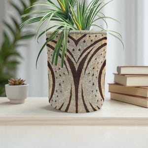 May include: A cylindrical plant pot with a brown and beige pattern, holding a green plant with long, thin leaves. The pot sits on a light-colored wooden surface. A small succulent in a white pot is to the left.
