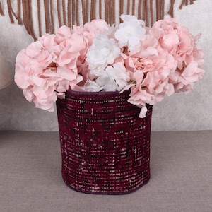 Puede incluir: Una cesta burdeos tejida llena de flores artificiales rosas y blancas. La cesta tiene forma cilíndrica y una superficie texturizada. Las flores están dispuestas en una exhibición completa y exuberante, creando un vibrante arreglo floral.