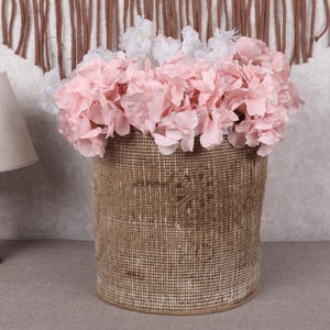 May include: A woven basket filled with pink and white flowers. The basket is cylindrical and has a textured, natural appearance. A wooden lamp with a beige lampshade is to the left. A decorative wall hanging is in the background.