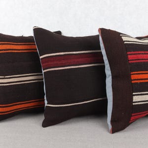 May include: Three decorative throw pillows with a dark brown background and horizontal stripes in orange, red, and cream. The pillows have a textured appearance, suggesting a woven fabric. The pillows are square-shaped and appear to be made of a durable material.