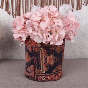 May include: A cylindrical vase made from a patterned rug holds a bouquet of pink and white flowers. The vase has a dark background with red, brown, and blue geometric designs. A wooden lamp with a beige lampshade is to the left.