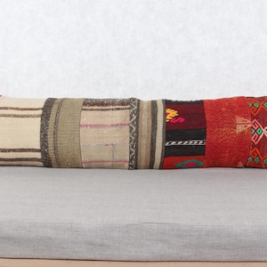 May include: A long, rectangular pillow with a patchwork design. The pillow features various fabric sections in shades of beige, brown, red, and orange. The pillow is placed on a gray surface, and the background is a plain white wall.