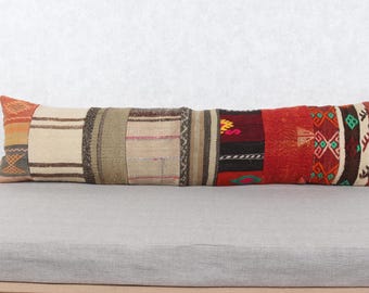 May include: A long, rectangular pillow with a patchwork design. The pillow features various fabric sections in shades of beige, brown, red, and orange. The pillow is placed on a gray surface, and the background is a plain white wall.