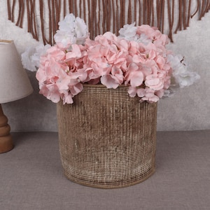 May include: A woven basket filled with pink and white flowers. A wooden lamp with a beige lampshade is to the left. The background features a textured wall and a decorative wall hanging.
