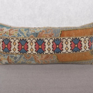 May include: Rectangular decorative pillow with a patterned design. The pillow features a central band with red, blue, and yellow geometric shapes on a cream background, framed by orange and blue sections. The pillow is placed on a gray surface.