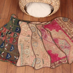 May include: A collection of colorful, patterned rugs in shades of pink, green, and beige, arranged in a fan shape on a wooden floor. A woven basket with a white cushion is in the background.