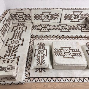 May include: An off-white sectional sofa with geometric brown patterns. The set includes a large rug, cushions, and a small ottoman. The design features a repeating tribal-inspired motif, creating a cohesive and stylish seating area.