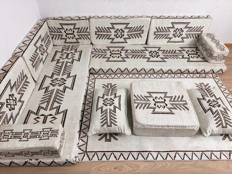 May include: An off-white sectional sofa with geometric brown patterns. The set includes a large rug, cushions, and a small ottoman. The design features a repeating tribal-inspired motif, creating a cohesive and stylish seating area.