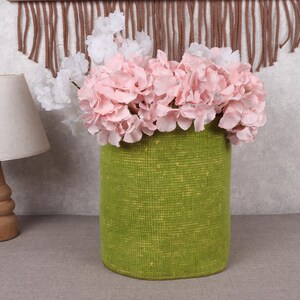 Può includere: Un vaso cilindrico verde pieno di fiori rosa e bianchi. Il vaso ha una superficie strutturata e poggia su una superficie grigia. A sinistra c'è una lampada in legno con un paralume beige.