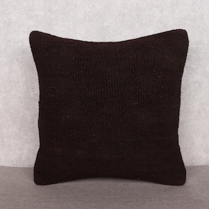 May include: A square, dark brown woven pillow. The pillow has a textured surface and a slightly curved top edge. The pillow is placed on a gray surface against a light gray background.