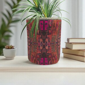 May include: A cylindrical plant pot with a colorful, patterned design in shades of red, brown, and pink. The pot contains a green plant with long, slender leaves. The pot is sitting on a light-colored wooden surface.