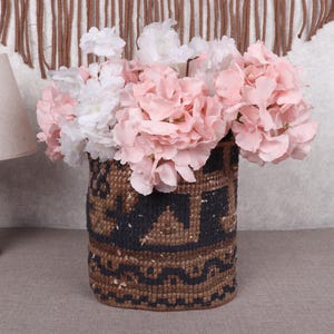 May include: A woven basket with a geometric pattern in brown and black, filled with pink and white artificial flowers. The basket is cylindrical and sits on a neutral surface.