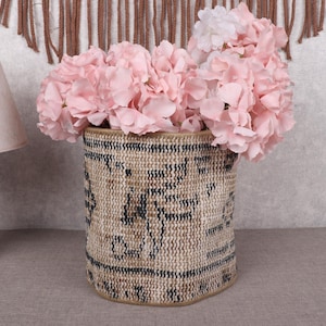 May include: A woven basket with a beige and black pattern, filled with pink flowers. The basket is cylindrical and sits on a gray surface. A wooden lamp with a beige lampshade is to the left.