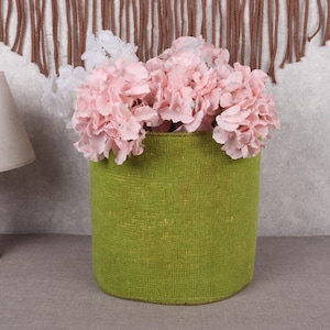 May include: A green woven basket filled with pink and white flowers. The cylindrical basket sits on a gray surface, with a wooden lamp and a decorative wall hanging in the background. The flowers are a focal point.