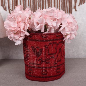 May include: A red cylindrical vase with a dark pattern holds a bouquet of light pink flowers. The vase is made of a woven material. A wooden lamp with a white lampshade is to the left.