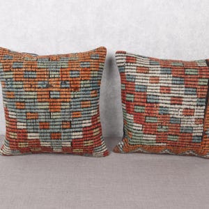 May include: Two square throw pillows with a woven pattern in shades of orange, red, blue, and white. The pillows have a textured appearance and are placed on a neutral surface.