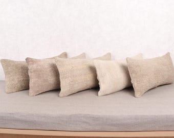 May include: Six rectangular throw pillows in varying shades of beige and tan are arranged on a light gray surface. The pillows have a textured, woven appearance, suggesting a natural fiber material. The background is a plain, off-white wall.