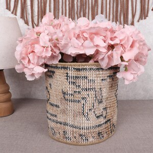 May include: A woven basket with a geometric pattern in black and tan holds a large arrangement of pink flowers. A wooden lamp with a beige lampshade sits to the left of the basket. The scene is set on a neutral-toned surface.