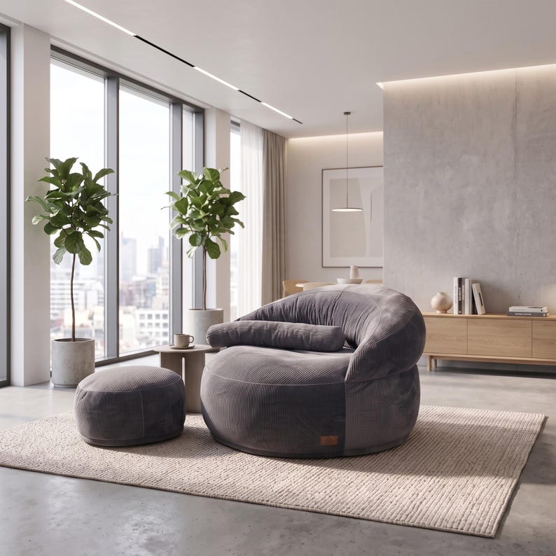 May include: A dark gray corduroy lounge chair with a rounded back and a matching cylindrical bolster pillow, accompanied by a circular corduroy ottoman. The furniture set is positioned on a textured area rug in a bright, open-concept room.
