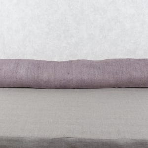 May include: A long, rectangular lavender-colored pillow. The pillow has a textured surface and is resting on a gray surface. The pillow is a decorative item, likely for a bed or sofa.
