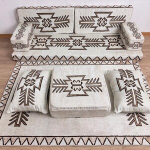 May include: A cream-colored floor seating set with geometric patterns in brown. The set includes a sofa, two armrests, a rug, a square ottoman, and two pillows. The rug and cushions have a repeating geometric design.