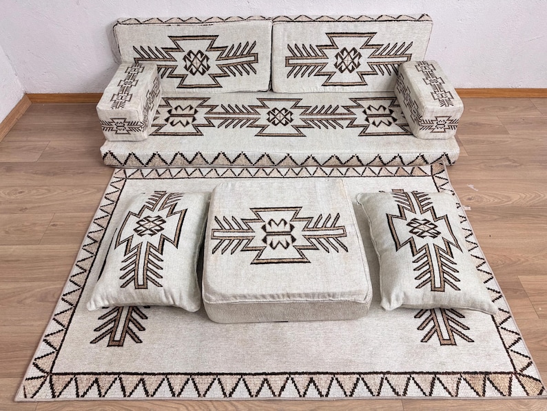 May include: A cream-colored floor seating set with geometric patterns in brown. The set includes a sofa, two armrests, a rug, a square ottoman, and two pillows. The rug and cushions have a repeating geometric design.