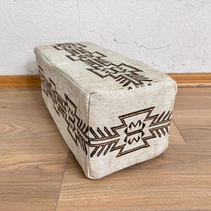 May include: A rectangular beige ottoman with a geometric brown pattern. The design is repeated on the top and sides of the ottoman. The ottoman is placed on a wooden floor.