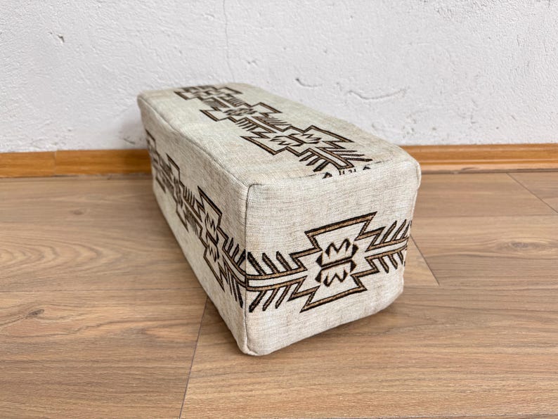 May include: A rectangular beige ottoman with a geometric brown pattern. The design is repeated on the top and sides of the ottoman. The ottoman is placed on a wooden floor.