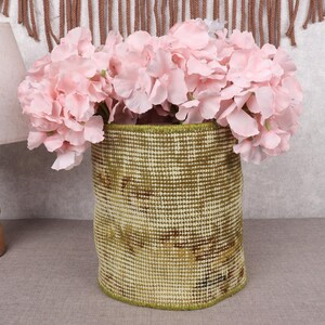 May include: A woven basket with a green and beige pattern holds a bouquet of pink flowers. The basket is cylindrical and has a green trim. A wooden lamp with a beige lampshade is to the left.