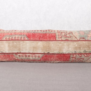 May include: Rectangular decorative pillow with a vintage rug design. The pillow features a central beige stripe bordered by red and faded floral patterns. The pillow is placed on a gray surface.