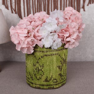 May include: A green woven vase filled with pink and white flowers. The vase has a textured pattern and sits on a gray surface. A wooden lamp and a wall hanging are in the background.
