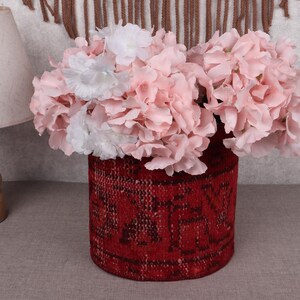 May include: A cylindrical, red woven vase filled with pink and white flowers. The vase has a patterned design. A wooden lamp with a beige lampshade is in the background.