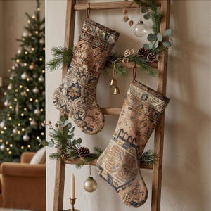 May include: Two Christmas stockings featuring a detailed, multi-colored woven pattern in shades of tan, blue, and cream. The stockings are hung on a wooden ladder decorated with pine branches, small pinecones, and hanging metallic ornaments.