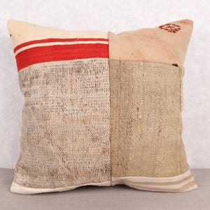 May include: Square decorative pillow with a patchwork design. The pillow features a cream-colored top with a red stripe, a tan woven section, and a light brown woven section. The pillow is a neutral color and would complement any decor.