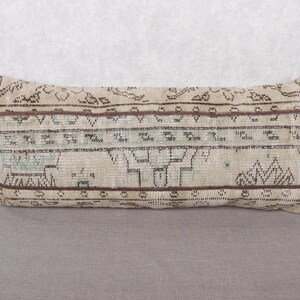 May include: Rectangular beige pillow with a geometric pattern in brown and teal. The pillow has a vintage, distressed look, with faded colors and a textured surface. The design features repeating shapes and lines.