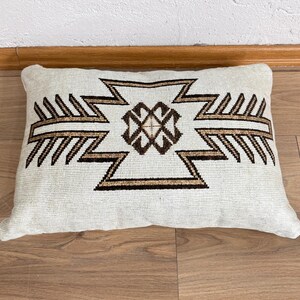 May include: Rectangular off-white pillow with a geometric design in brown and black. The pattern features a central diamond shape with zigzag and arrow-like elements. The pillow is placed on a wooden surface.