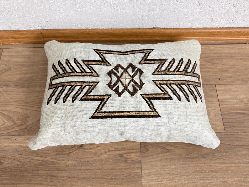 May include: Rectangular off-white pillow with a geometric design in brown and black. The pattern features a central diamond shape with zigzag and arrow-like elements. The pillow is placed on a wooden surface.