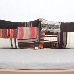May include: Two rectangular throw pillows with patchwork designs. The pillows feature a mix of colors including brown, black, red, orange, and cream. The pillows are placed on a light gray surface against a white wall.