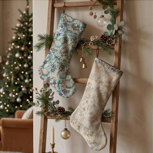 May include: Two holiday stockings hanging on a wooden ladder. The left stocking features a cream base with a dense floral pattern in teal, blue, and pink. The right stocking has a muted beige and grey vintage-style ornate pattern.