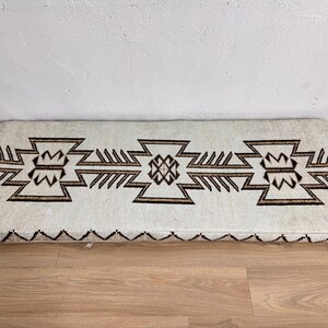 May include: Rectangular off-white rug with a geometric brown pattern. The design features repeating stylized shapes and a zig-zag border. The rug is placed on a wooden floor.