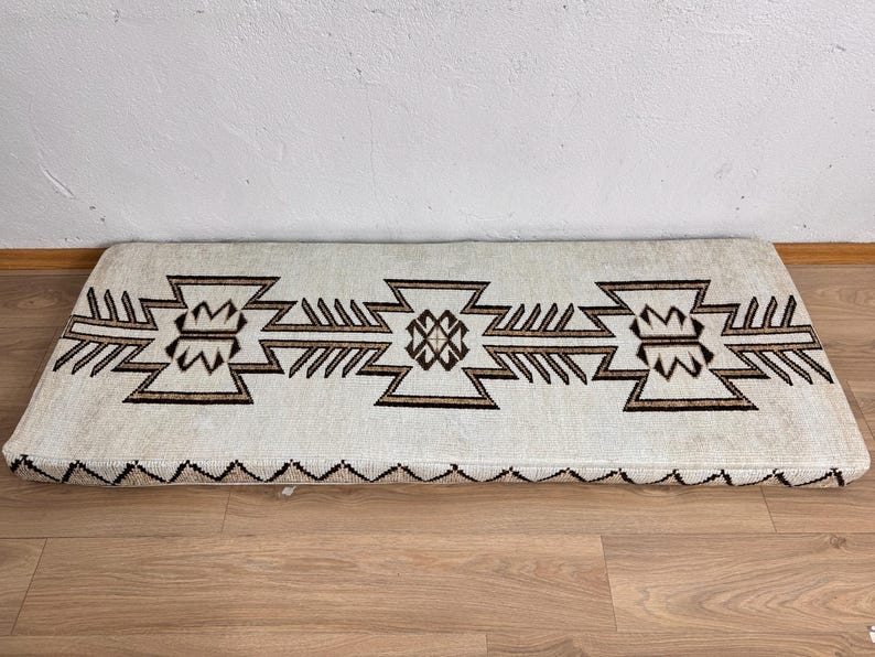 May include: Rectangular off-white rug with a geometric brown pattern. The design features repeating stylized shapes and a zig-zag border. The rug is placed on a wooden floor.