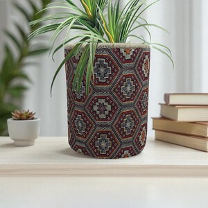 May include: A cylindrical plant pot with a geometric pattern in shades of red, black, and beige. The pot holds a green plant with long, slender leaves. The pot sits on a light-colored wooden surface.
