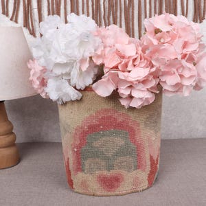 May include: A woven basket with a colorful design holds a bouquet of pink and white flowers. A wooden lamp with a beige lampshade is to the left of the basket. The background is a neutral color.