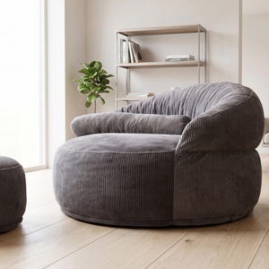 May include: A round, low-profile lounge chair upholstered in dark gray corduroy fabric. The chair features a deep, curved backrest and a wide, plush seat cushion, accompanied by a matching horizontal bolster pillow across the center.