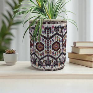 Könnte beinhalten: Ein zylindrischer Blumentopf mit einem farbenfrohen geometrischen Muster, der eine grüne Pflanze mit langen, schlanken Blättern enthält. Das Design des Topfes zeigt sich wiederholende Formen in Rot, Blau, Gelb und anderen Farben. Der Topf steht auf einer hellen Holzoberfläche.