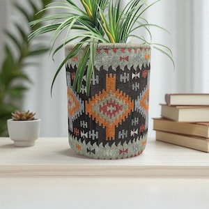 Op de afbeelding: Een cilindrische plantenpot met een geometrisch patroon in bruin-, oranje- en grijstinten. De pot bevat een groene plant met lange, slanke bladeren. De pot staat op een lichtgekleurd houten oppervlak.